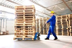 Warehouse worker in blue coveralls and yellow hard hat moving a tall stack of wood pallets with a pallet jack inside a pallet manufacturer facility